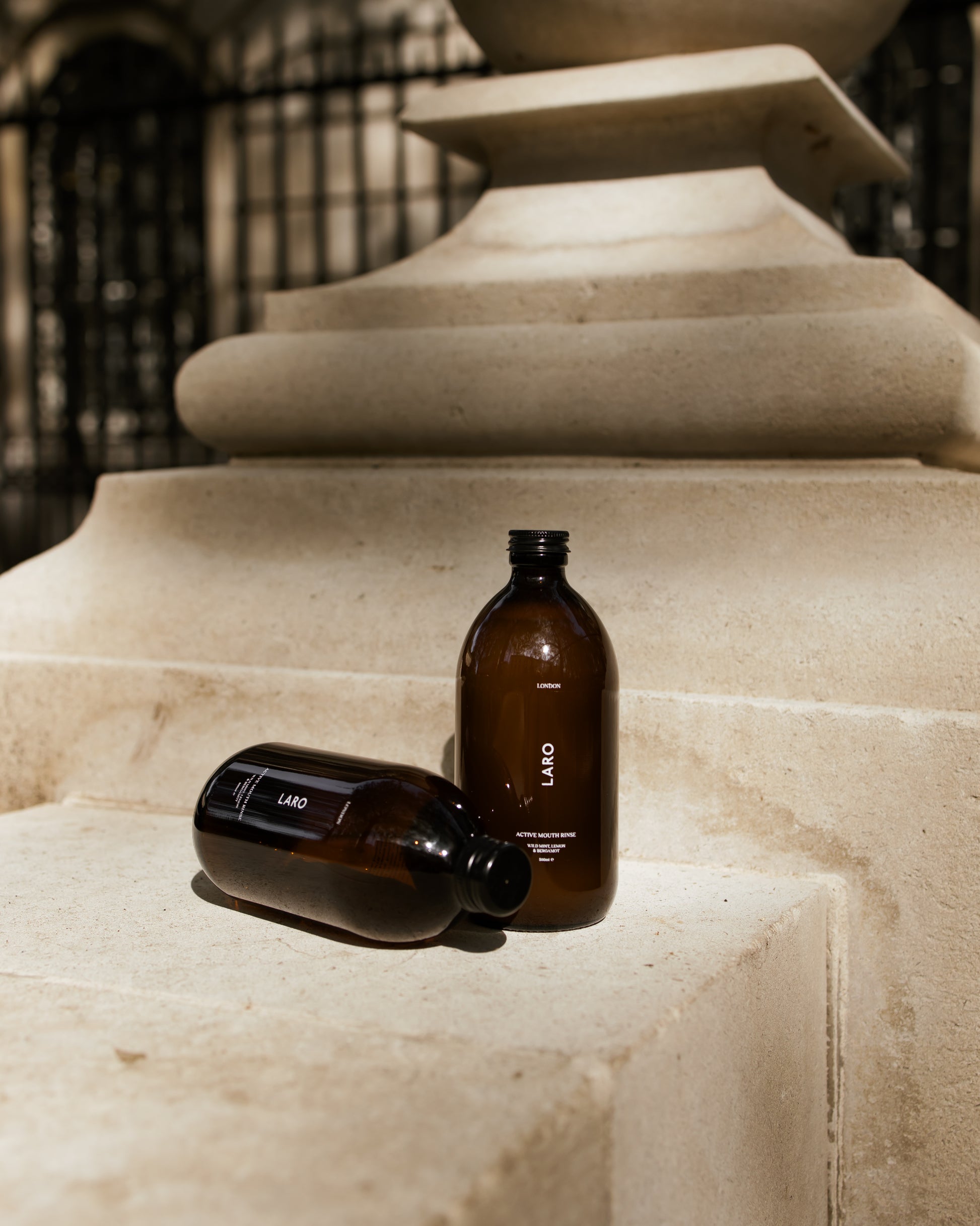2 dark amber glass Laro Active Mouth Rinse bottles with black aluminium caps, one standing one lying down, set against a Buckingham palace stone wall in London. .
