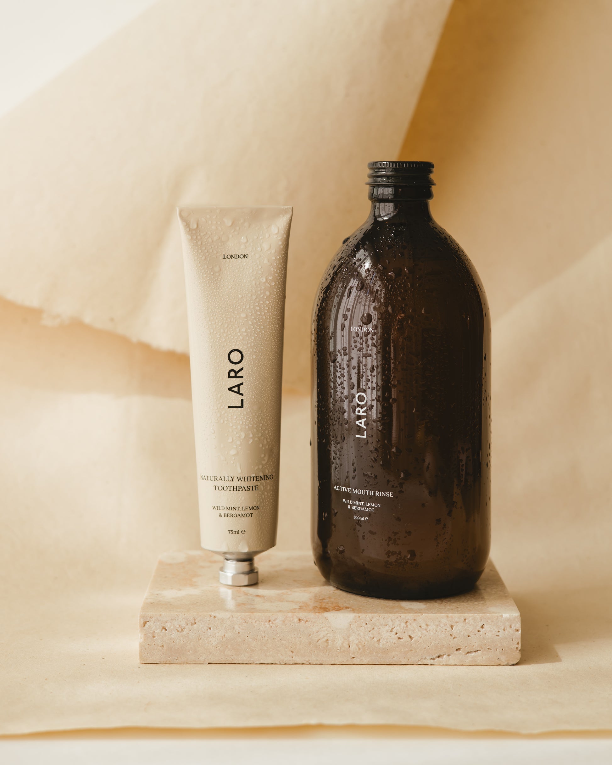 Laro's signature range - Naturally Whitening Toothpaste aluminium off-white tube, with Laro's dark amber glass Active Mouth Rinse bottle both in wild mint, lemon and bergamot flavour. Stood next to each other with condensation on the product on a marble tile and paper background.