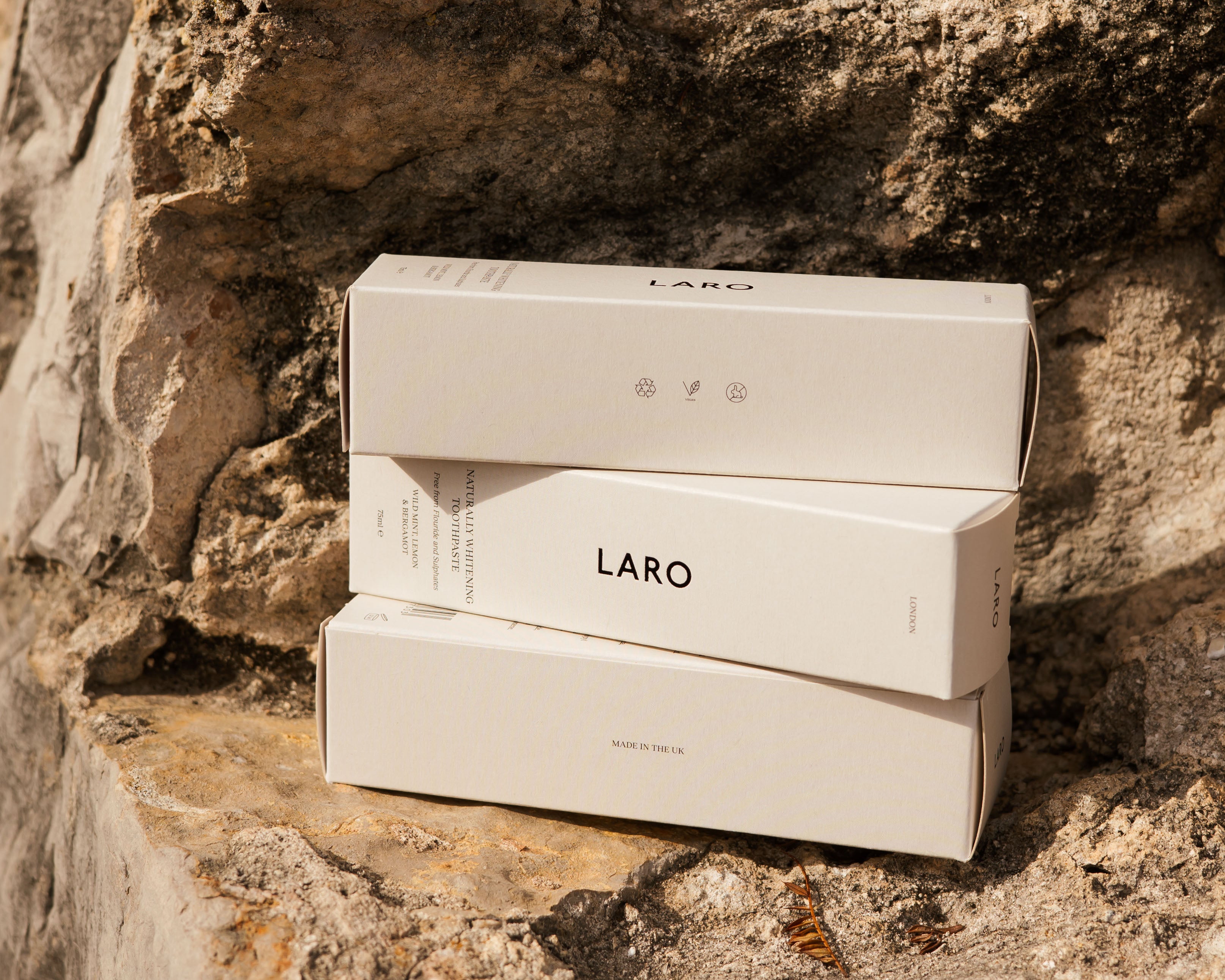 Stack of three product boxes for Laro's Naturally Whitening Toothpaste, SLS free and fluoride free, stacked to show the recyclable, vegan and cruelty free symbols as well as the Made in the UK stamp. Set against a stone wall backdrop.