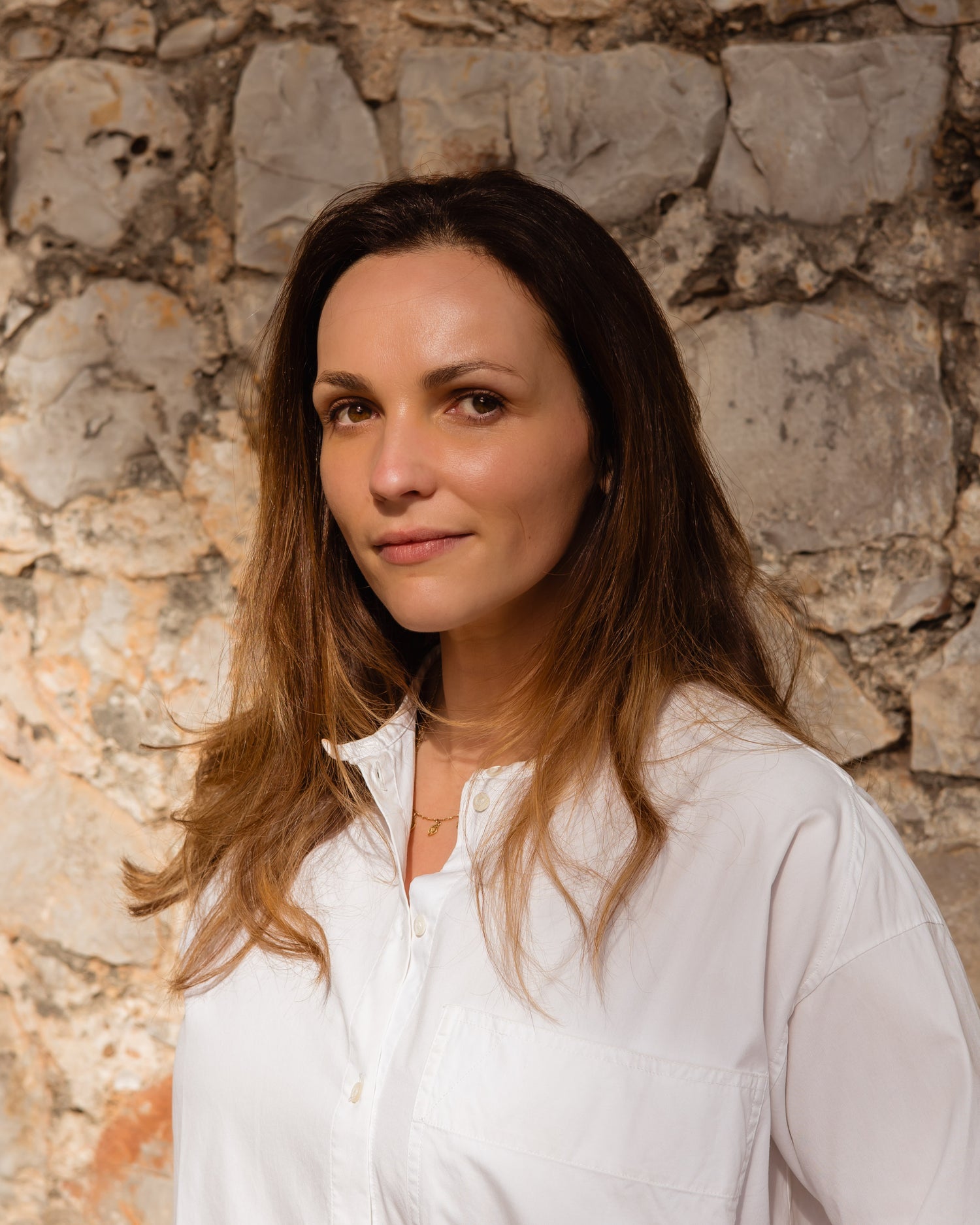 Head shot of founder, white skin and long brown hair wearing a white shirt, stone-wall background.