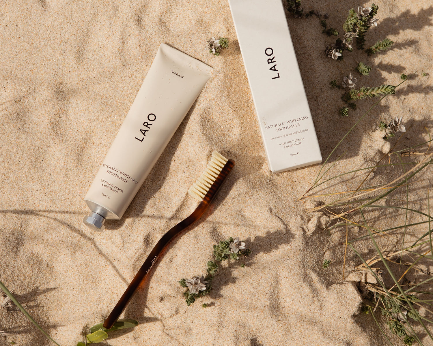 Beach landscape with a Laro aluminium Naturally Whitening Toothpaste tube, a packaging box for the toothpaste and a brown cellulose acetate toothbrush lying in the sand, with beach green grass and flower shoots surrounding. 